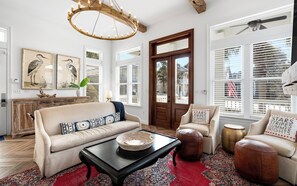 A living room with a sofa, armchairs, a coffee table, and a chandelier. There are framed pictures on the wall, a wooden cabinet, and a door with a view outside.