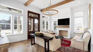Un vaste salon doté d’un parquet en bois disposé en point de Hongrie, d’une cheminée, d’un grand téléviseur fixé au-dessus et d’un ventilateur de plafond.