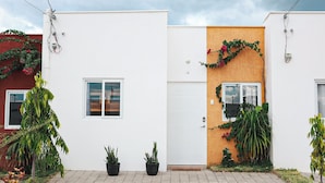Una casa moderna de dos pisos con fachada blanca, una pared texturizada de madera a la derecha y un pequeño jardín con plantas en macetas y un sendero.