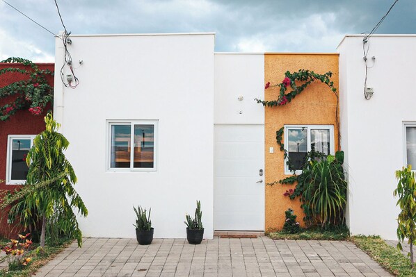 Ett modernt hus i två våningar med en vit fasad, en vägg med trästruktur på höger sida och en liten trädgårdsyta med planterade blommor och en gångstig.