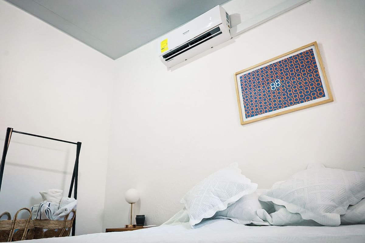 Una habitación de hotel con cama, aire acondicionado de pared, un cuadro con un diseño geométrico, una cesta de mimbre con toallas y una lámpara de noche.