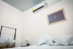 Ett hotellrum med en säng, en väggmonterad luftkonditioneringsanläggning, en inramad geometrisk mönsterbild, en korg av rotting med handdukar och en sänglampa.