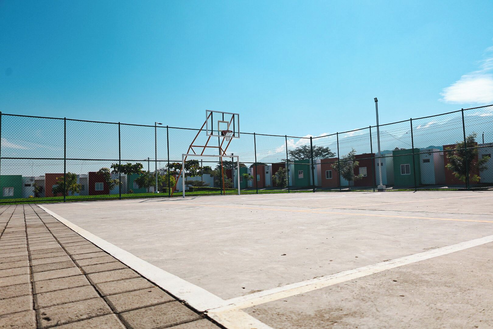 Un campo da basket all'aperto con un canestro in metallo, circondato da una recinzione in rete metallica.