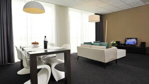 Una habitación de hotel moderna con comedor, sofá, televisión y un amplio ventanal con cortinas.