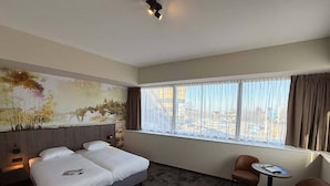 Habitación de hotel con una cama grande, una mesita pequeña, dos sillas, un cuadro en la pared y una ventana con cortinas.