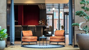 A lobby with two orange armchairs, a small table, a large potted plant, and a view of a brick building through large windows. - Stylish Forest-Edge Home — Convenient Trail Access, Kitchenette, & Free Parking (Oisterwijk)