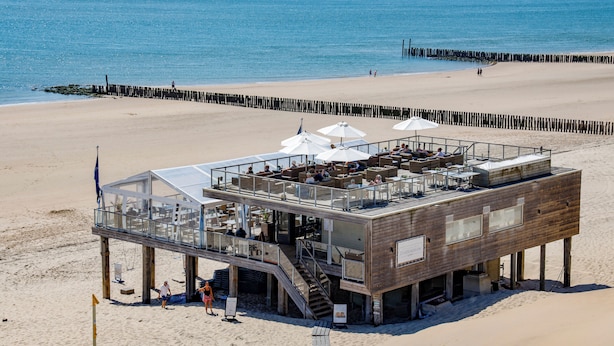 Una estructura de madera frente a la playa con asientos al aire libre y un cerramiento de cristal, posiblemente un restaurante o bar, con vista al mar.