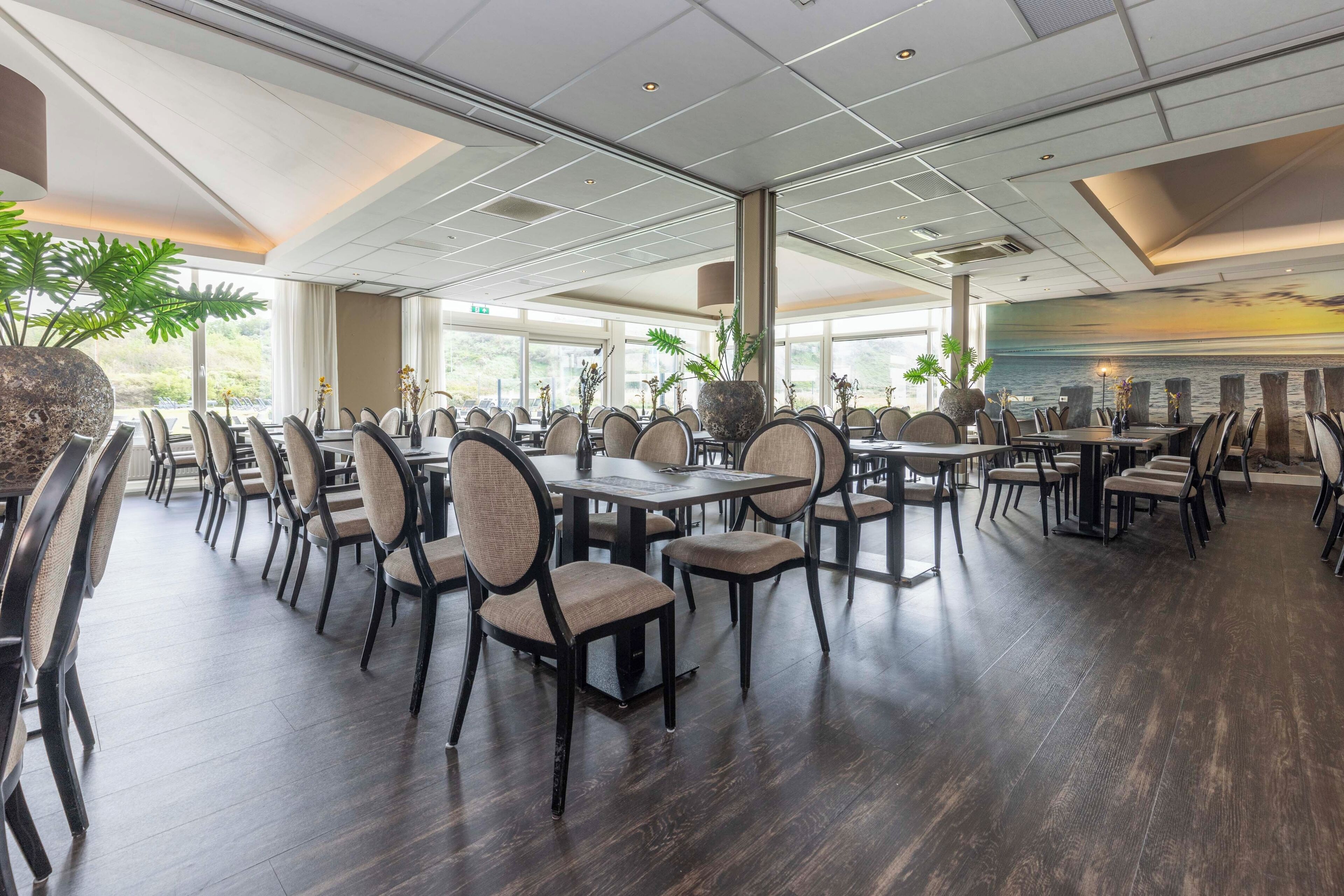 A spacious dining area with multiple tables and chairs, large windows, and indoor plants.