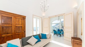 A modern living room with a sofa, a dining area with blue chairs, and a view of a mountainous landscape through large windows. - Villa Domi by My Villa Collection (Amalfi)