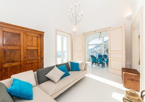 A modern living room with a sofa, a dining area with blue chairs, and a view of a mountainous landscape through large windows. - Villa Domi by My Villa Collection (Amalfi)