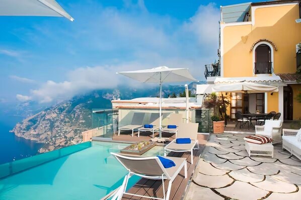A rooftop infinity pool with lounge chairs and umbrellas, overlooking a mountainous coastline. - Villa Deus by My Villa Collection (Nocelle)