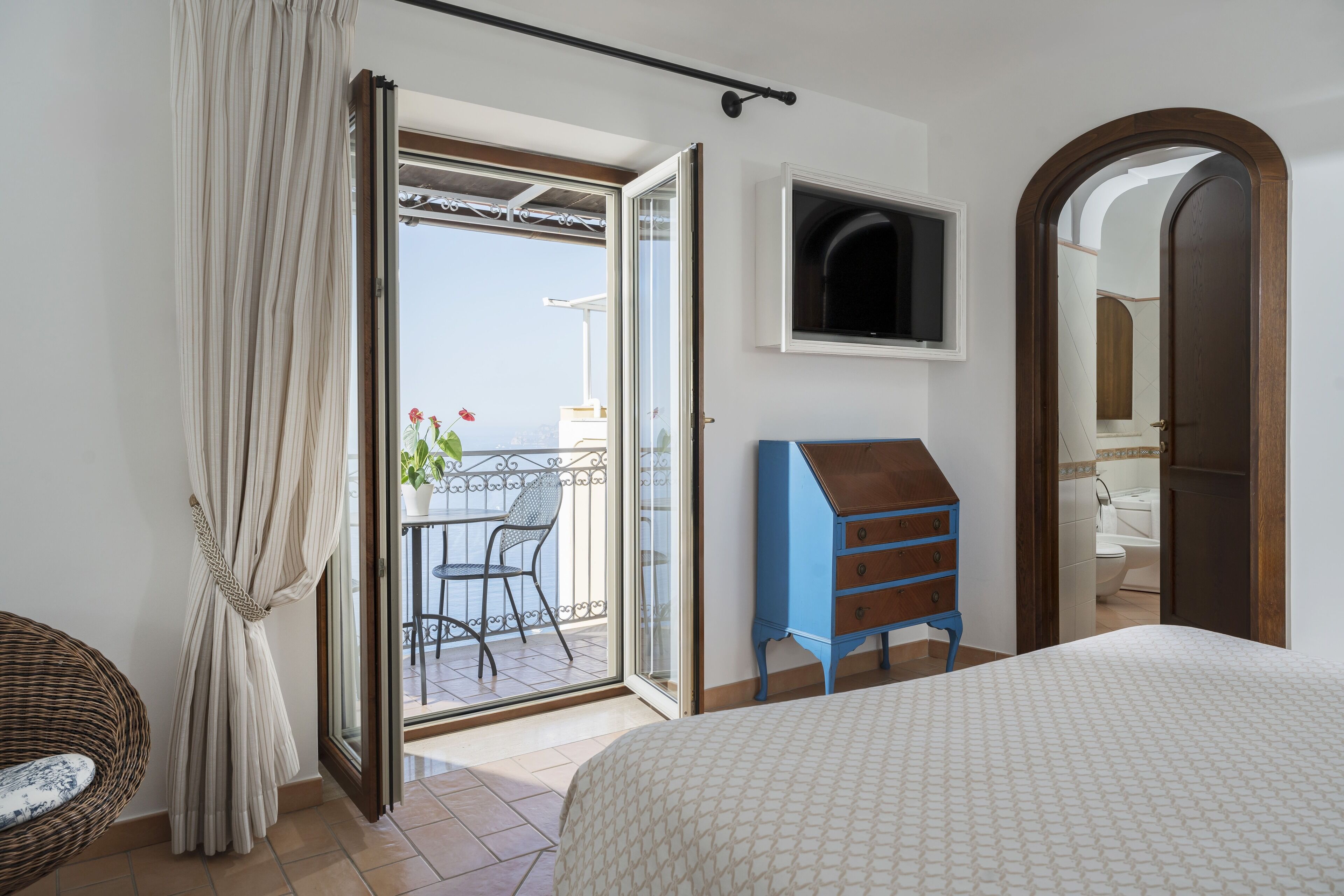 A bedroom with a bed, a dresser, a TV, and a balcony with a chair and potted plants.