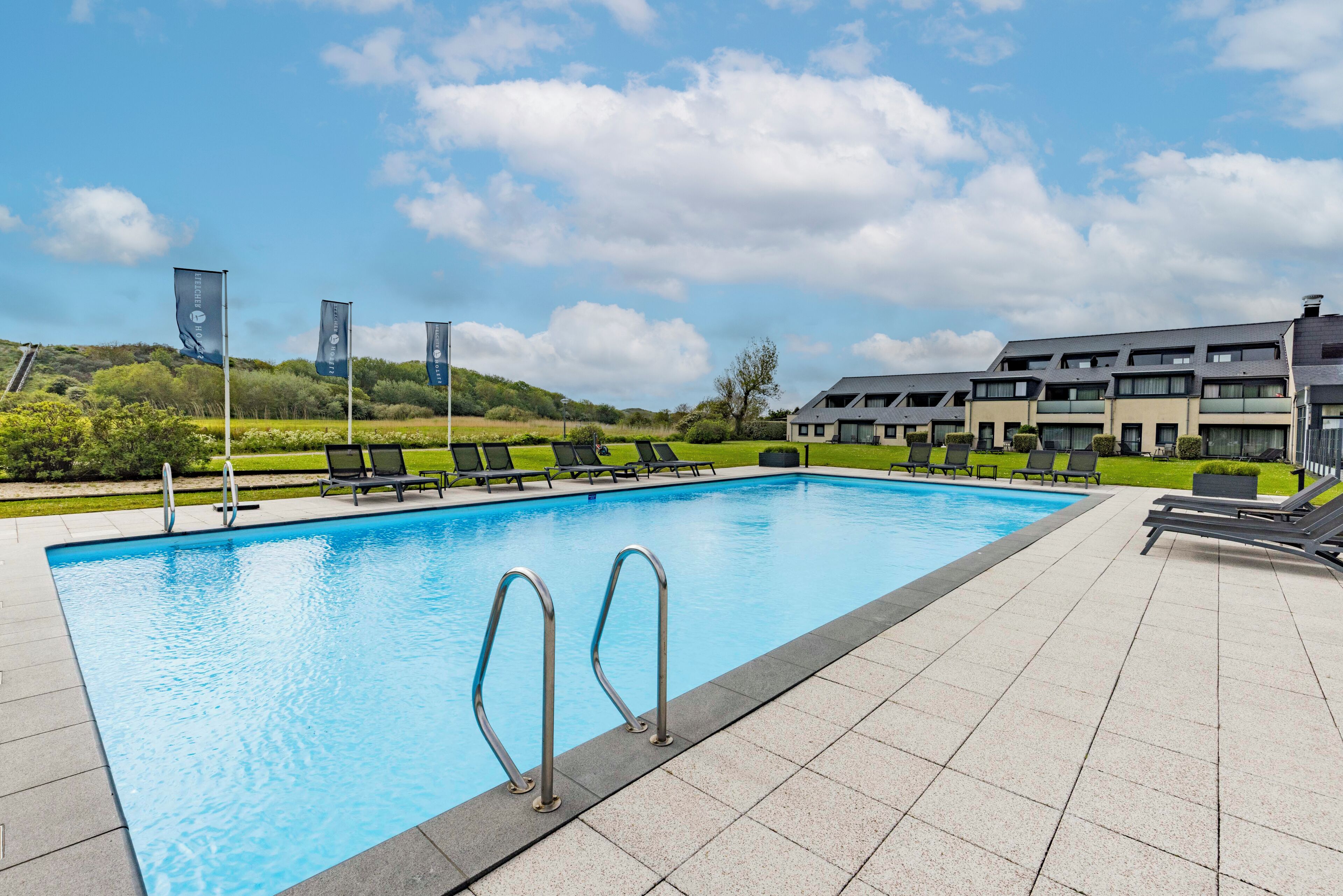 A swimming pool with metal ladders, surrounded by lounge chairs and a building with balconies.