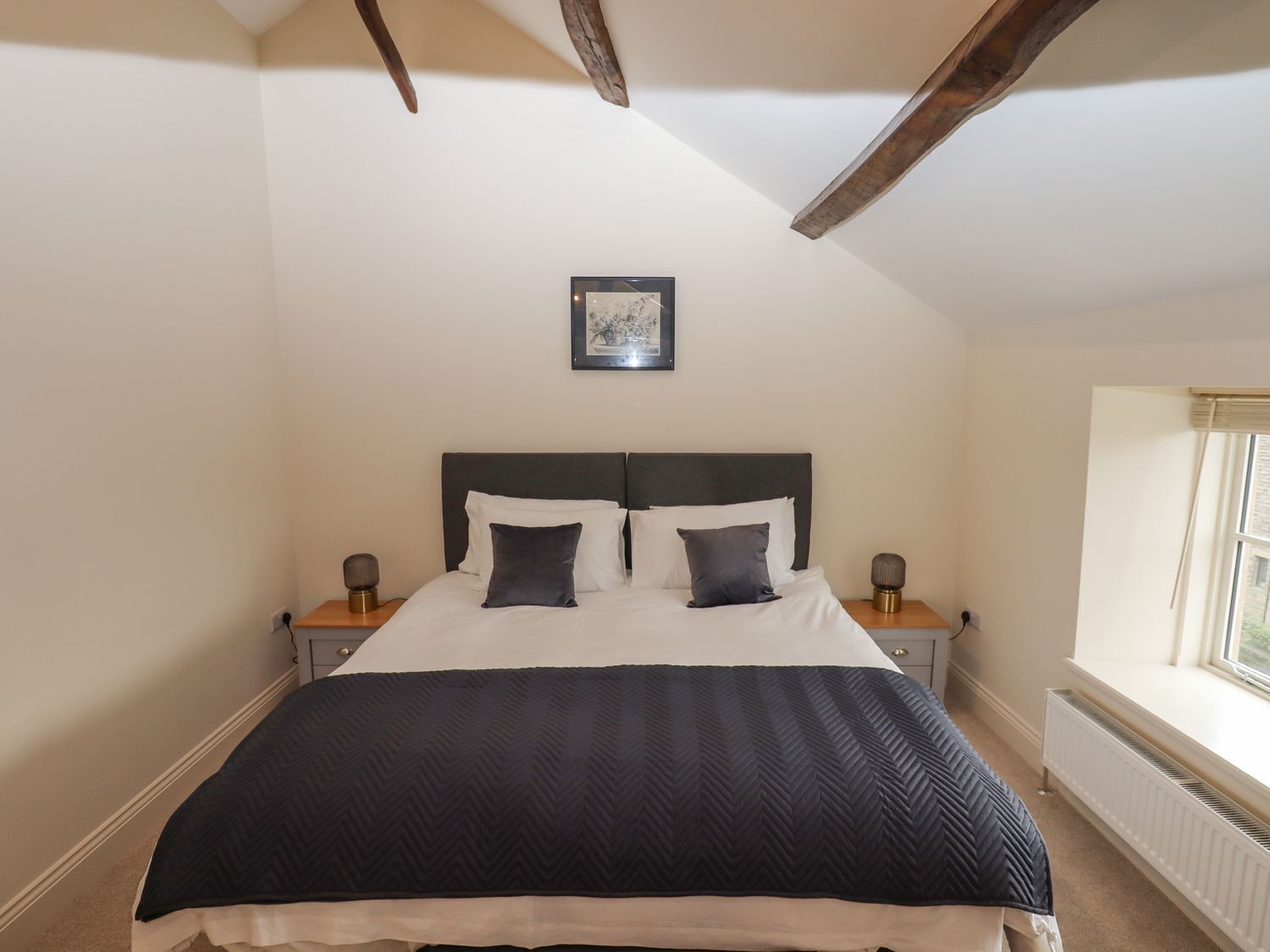 A bedroom with a large bed, two bedside tables, a framed picture on the wall, and a window with a view.