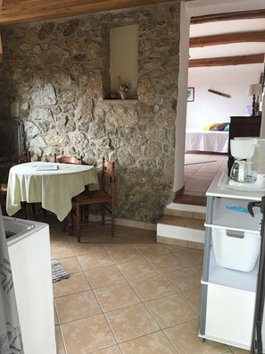 Ein Zimmer mit Steinwänden, einem Esstisch mit Stühlen, einer kleinen Küchenzeile mit Spüle und einem Bett im Hintergrund.