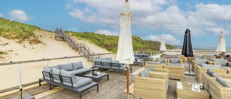 Eine Holzterrasse mit Sitzgelegenheiten im Freien, darunter Sofas und Stühle, mit Blick auf einen Strand.