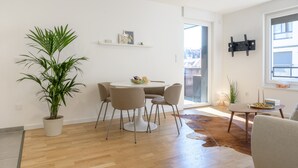 A modern living room with a dining table, chairs, a potted plant, a small table with candles, and a mounted TV. - Colmar Chic – Modern Old Town Apartment with Wi-Fi, Garage, Elevator for 2–6 Guests (Colmar)