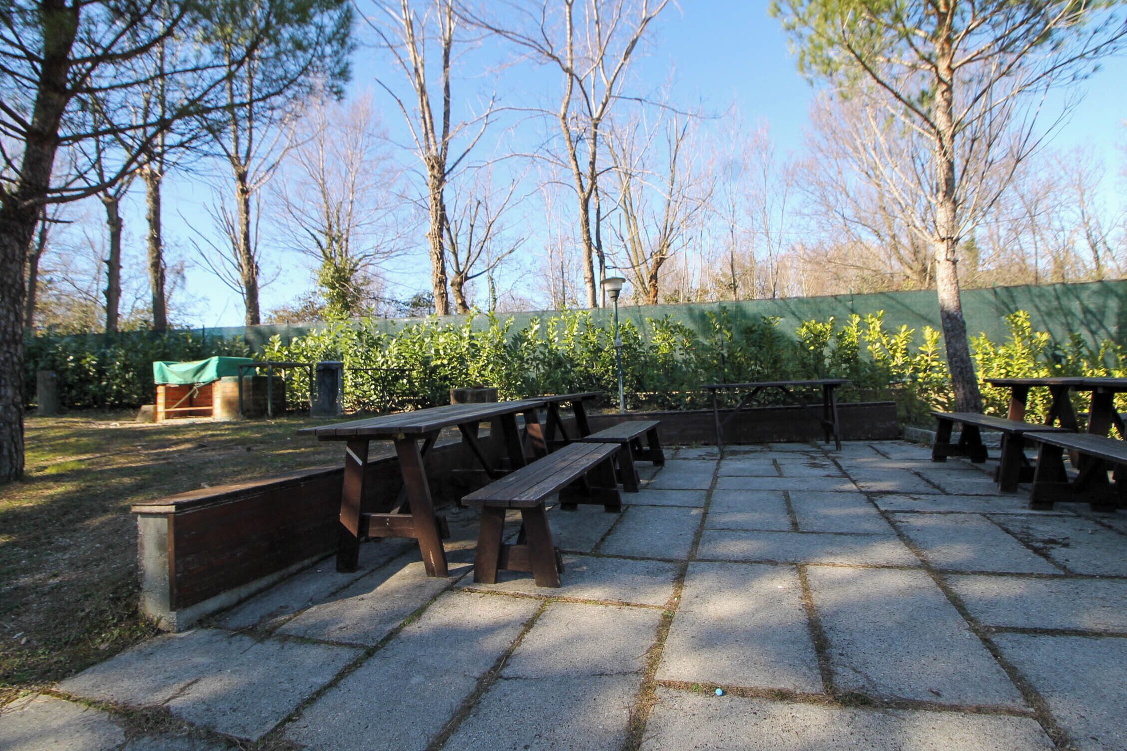 Un'area esterna con tavoli da picnic disposti su un patio in pietra, circondata da alberi e un recinto verde.
