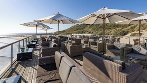 A rooftop bar with wicker furniture, outdoor umbrellas, and a view of the beach. - Beachfront Escape with Direct Shore Access, Pool & Sunset Terrace Views (Westkapelle)