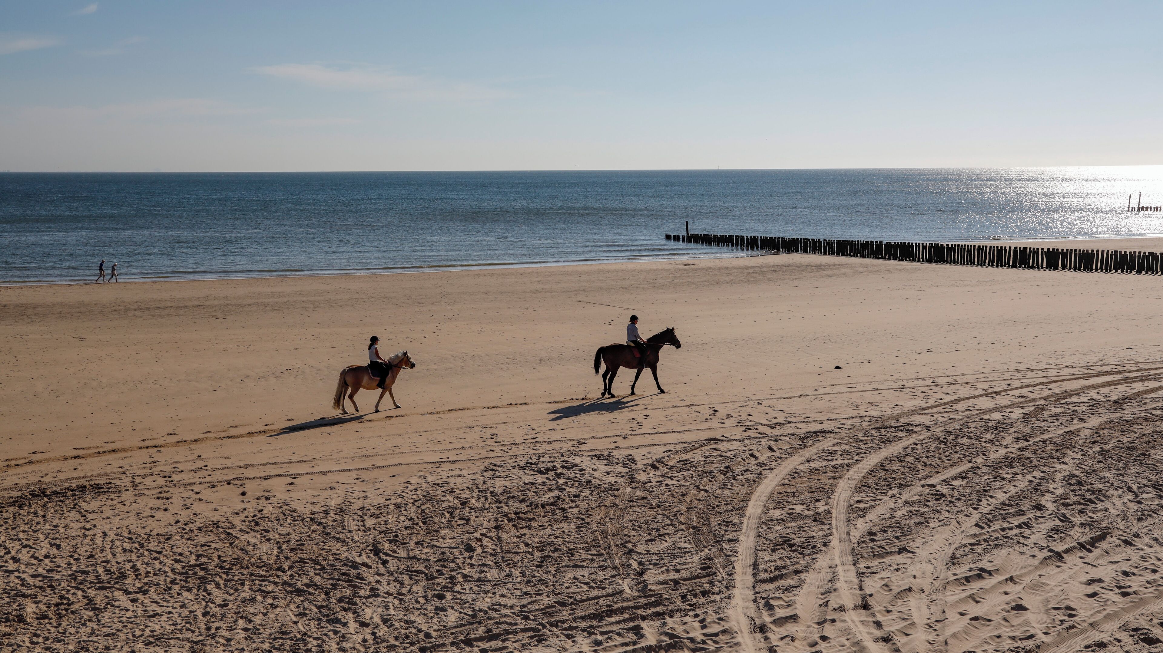 A beach with two horses and riders, a wooden pier, and a clear sky.