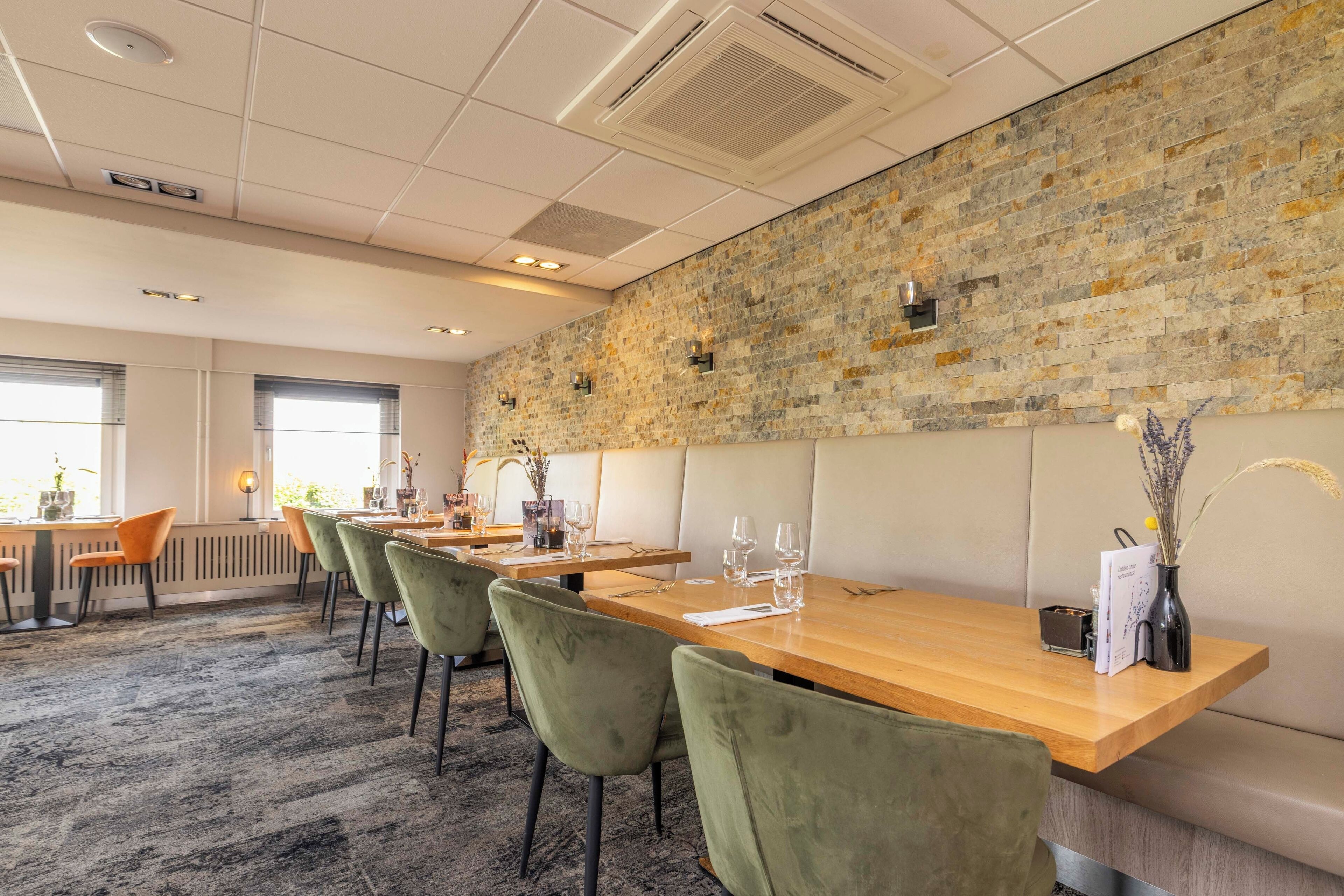 A restaurant with wooden tables and green chairs, a stone wall, and a window with natural light.