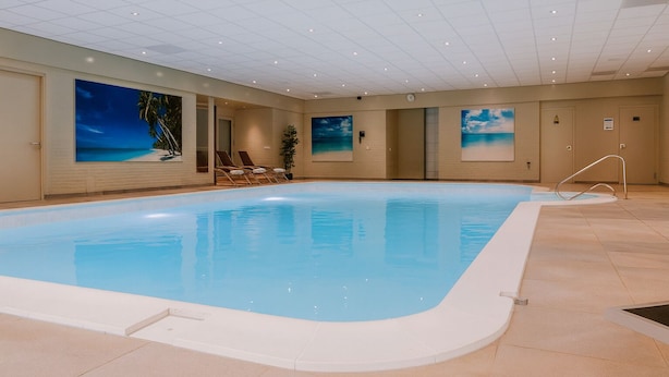 Una piscina interior con agua azul cristalina, rodeada de paredes color beige y un suelo de baldosas. Hay sillones de descanso a lo largo de la piscina y un cuadro grande con una escena de playa en la pared.