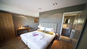 Habitación de hotel con una cama grande, suelo de madera, un escritorio con silla, un armario y un baño visible a través de una puerta abierta.