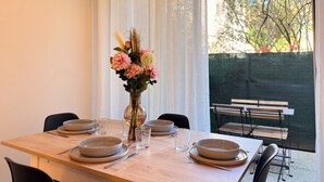 A dining table set with plates, bowls, and cutlery, a vase with flowers, and a view of an outdoor area with a bench. - Le Cosy du lagon Bleu (Fréjus)