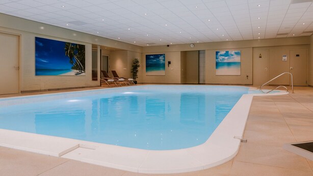 Une piscine intérieure à l’eau d’un bleu limpide, entourée de murs de couleur beige et dotée d’un sol carrelé. Des chaises longues sont disposées au bord de la piscine, et un grand tableau représentant une scène de plage orne le mur.