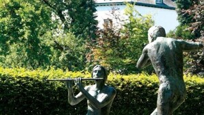 Un jardín de esculturas con tres estatuas, un banco de piedra y flores rojas.