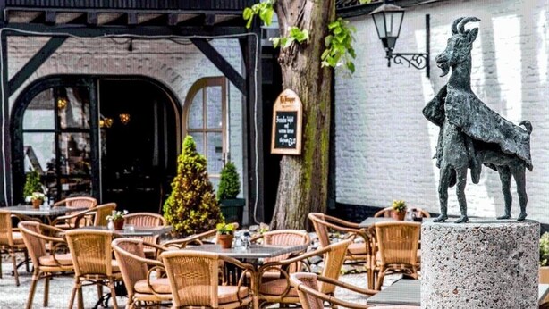 An outdoor cafe with wicker chairs and tables, a statue, and a building with a black awning and a signboard. - Peaceful Stay in Thorn – Explore Cobblestone Streets, Historic Buildings (Thorn)