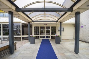 A covered entrance with a blue carpet leading to glass doors, benches, and planters.
