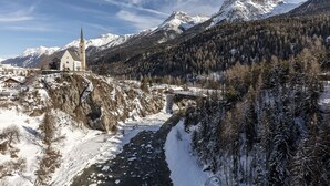 Una chiesa con un campanile slanciato, un ponte e montagne innevate sullo sfondo.