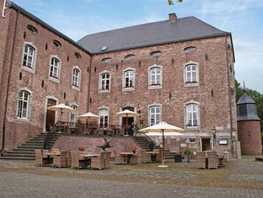 Ein großes Backsteingebäude mit zahlreichen Fenstern und einer Treppe, die zu einem Balkon führt. Draußen stehen Tische und Stühle mit Sonnenschirmen, und zur Rechten ist eine kleine, turmartige Struktur zu sehen.