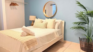 Una cama bien hecha con una toalla enrollada, una lámpara de mesita de noche, un aire acondicionado de pared, un espejo y una planta en maceta.