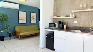 Cocina compacta con microondas, cafetera y lavabo. Hay una pequeña zona de comedor con una mesa y un sofá.