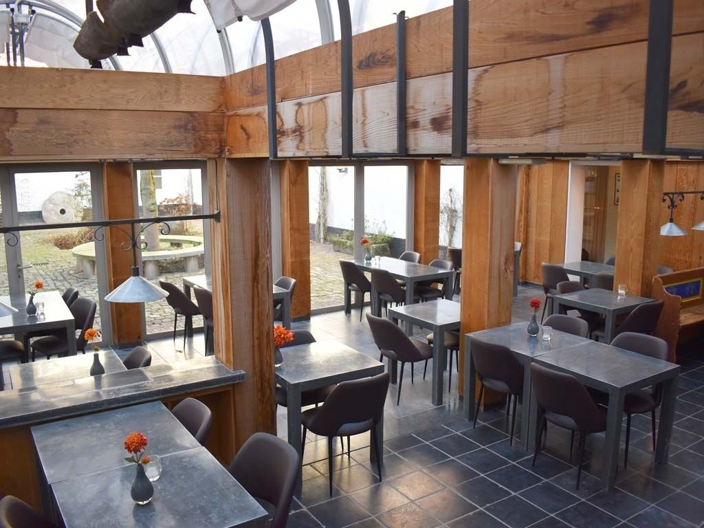 Een eethoek binnen met tafels en stoelen, grote ramen en uitzicht op het buitenleven.