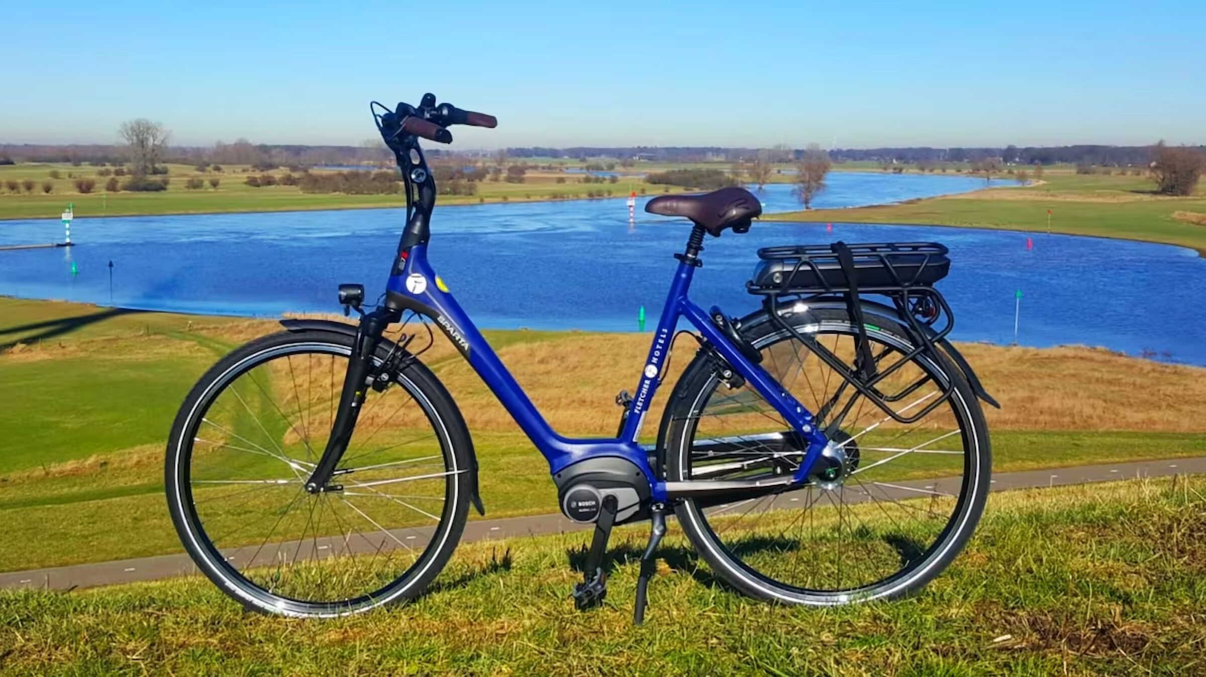 Ein blaues Fahrrad mit Korb und Gepäckträger, abgestellt im Gras in der Nähe von Wasser.