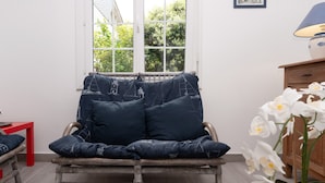 A wicker chair with dark cushions, a red table, and a wooden cabinet with a lamp. - Renovated Apartment with Private Garden, 150m from Saint-Colomban Beach (Carnac)