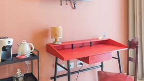 Una stanza con una TV a schermo piatto montata al muro, una scrivania rossa con una lampada, una sedia e uno scaffale con un bollitore e una bottiglia.