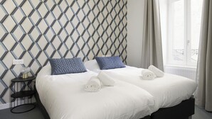 Camera d'albergo con parete a motivi geometrici, un letto con lenzuola bianche e due cuscini, un comodino con una lampada e una finestra con tende.