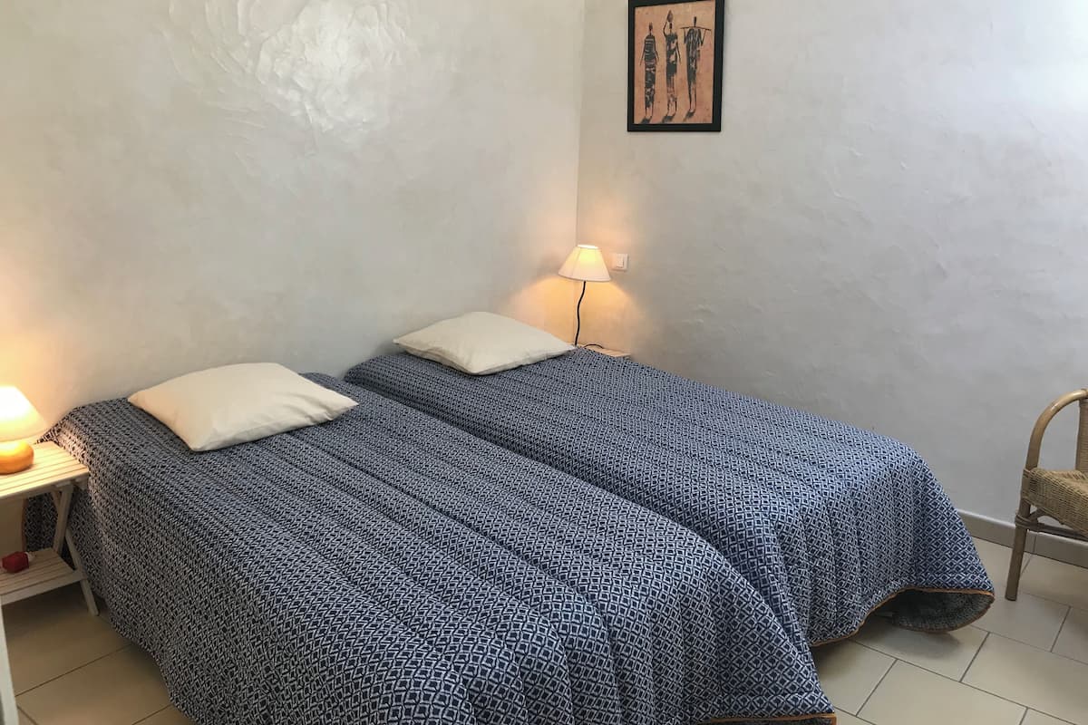A small, simple room with two beds, each with a patterned bedspread and a single pillow. There is a nightstand with a lamp and a framed picture on the wall.