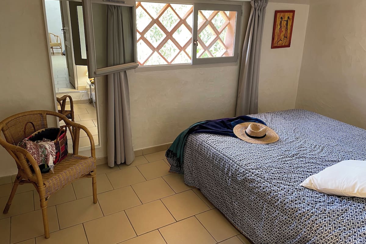 A bedroom with a bed, a wicker chair, a window with curtains, and a door leading to another room.