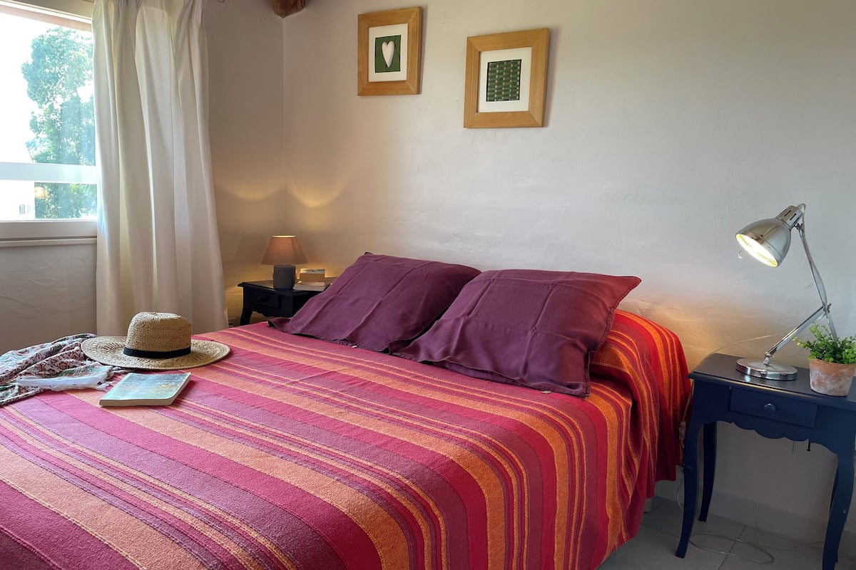 A neatly made bed with a striped bedspread, a hat, and a book on the left side. There are two framed pictures on the wall above the bed.