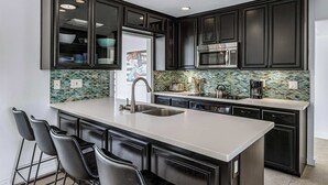Une cuisine moderne avec des meubles de couleur sombre, un grand îlot central doté d’un évier et un revêtement mural en carreaux.