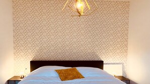 Habitación de hotel con papel tapiz estampado, cama con ropa blanca y una sola almohada amarilla, candelabro moderno y lámparas de noche.