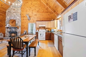 A cabin interior with a stone fireplace, wooden dining table, and a kitchen area with white appliances. - Juniper Presents: 10 Acre County Line Cabin - Private Pond & Firepit (Watervliet)