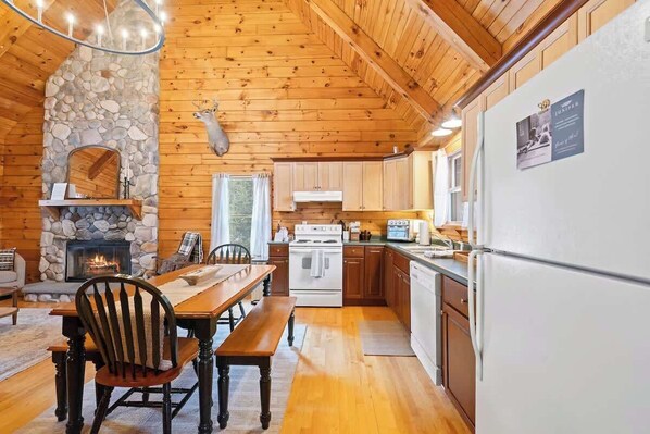 A cabin interior with a stone fireplace, wooden dining table, and a kitchen area with white appliances.