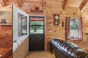 A wooden cabin interior with a door, a bench, a wall clock, and a window with curtains.