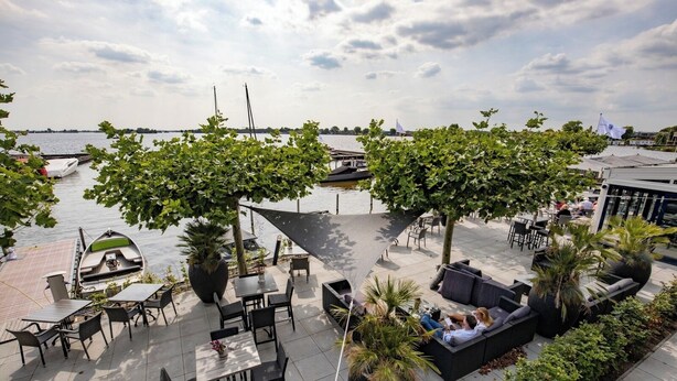 An outdoor seating area with tables and chairs, large planters, and a view of the water with boats.